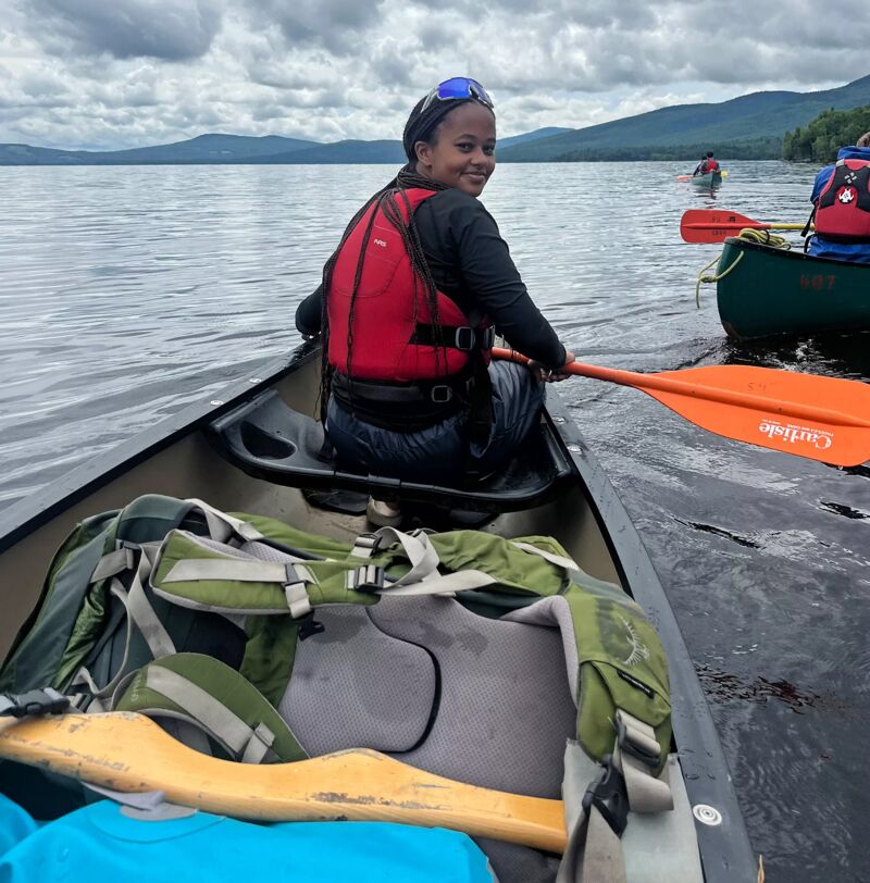 A young woman is paddling a canoe on a lake. She is wearing a red life jacket and has a paddle in her hand. There are other people in canoes in the background. The sky is cloudy and the water is calm. There is a green backpack in the canoe.